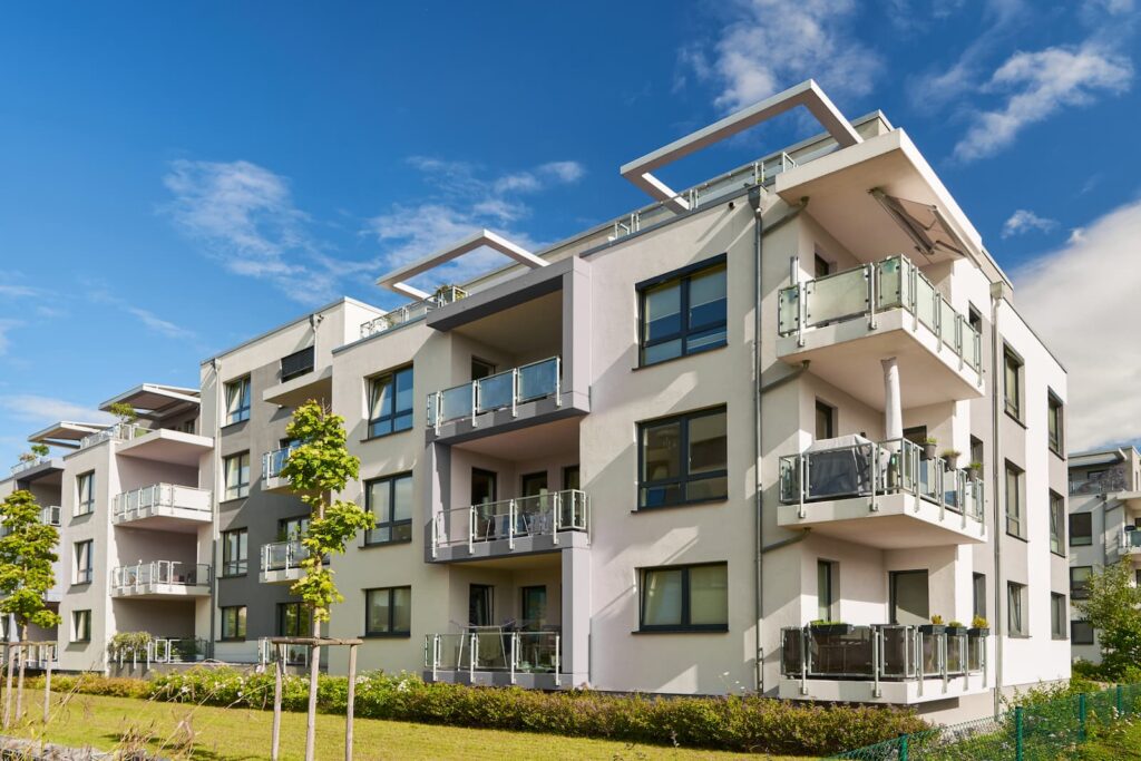 A bright, wide-angle exterior shot of a modern four-story apartment building with white and gray facades. The building features numerous balconies with glass railings and large windows, situated behind a manicured green lawn under a sunny blue sky.