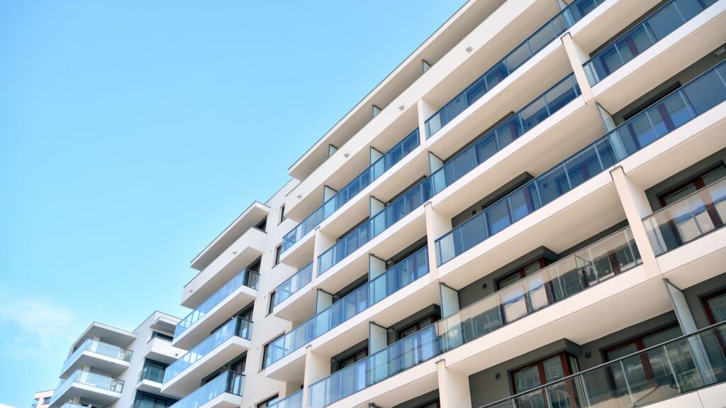 A low-angle upward view of a modern multi-story apartment building with white exterior walls. Multiple rows of balconies with clear glass railings and dark frames are stacked uniformly against a bright blue sky background.